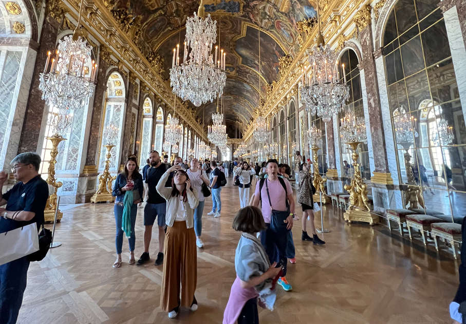 Turistas visitan la majestuosa Galería de los Espejos del Palacio de Versalles, admirando las grandes lámparas de cristal y el techo pintado.