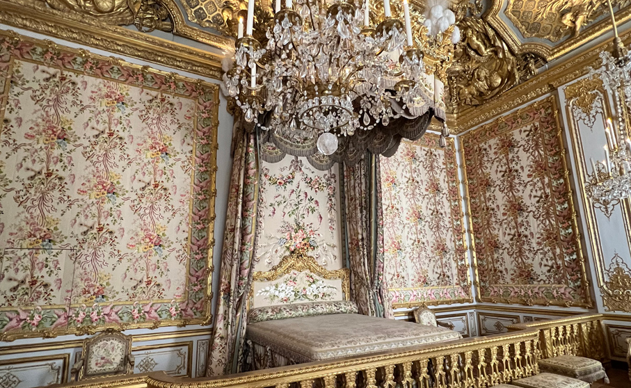 El suntuoso dormitorio de la Reina en el Palacio de Versalles, ricamente decorado con dorados, tapices florales y una gran lámpara de cristal.