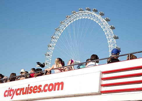 Des touristes à bord d'un bateau City Cruises sur la Tamise à Londres, avec la grande roue du London Eye en arrière-plan sous un ciel bleu.