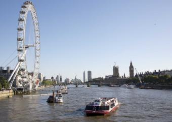 Panoramic view of the River Thames in London with the London Eye Ferris wheel on the left, a cruise boat in the center, and the Big Ben tower in the distance.