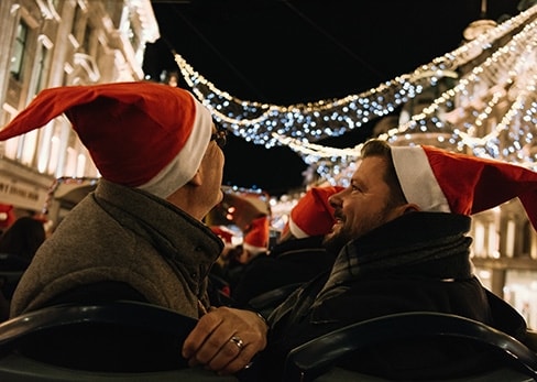Una pareja sonriente con gorros de Papá Noel en el piso superior de un Tootbus, admirando las guirnaldas de luces navideñas en Londres.