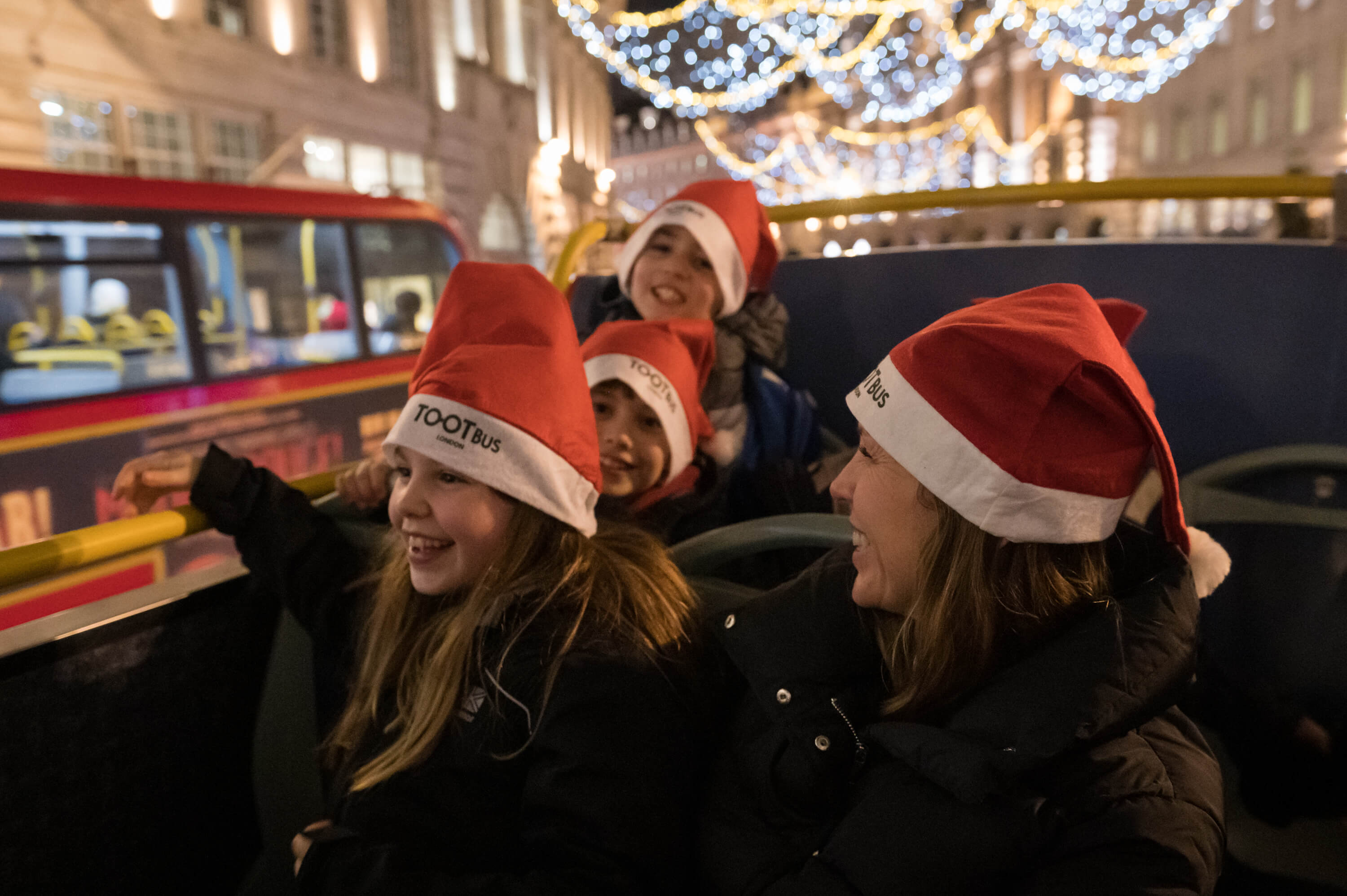 Una madre y sus tres hijos sonrientes con gorros de Papá Noel de Tootbus en el piso superior de un autobús descapotable durante un recorrido navideño por Londres.