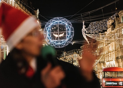 Un guía turístico desenfocado con un gorro de Papá Noel y un micrófono, con las luces navideñas de Regent Street al fondo en Londres.