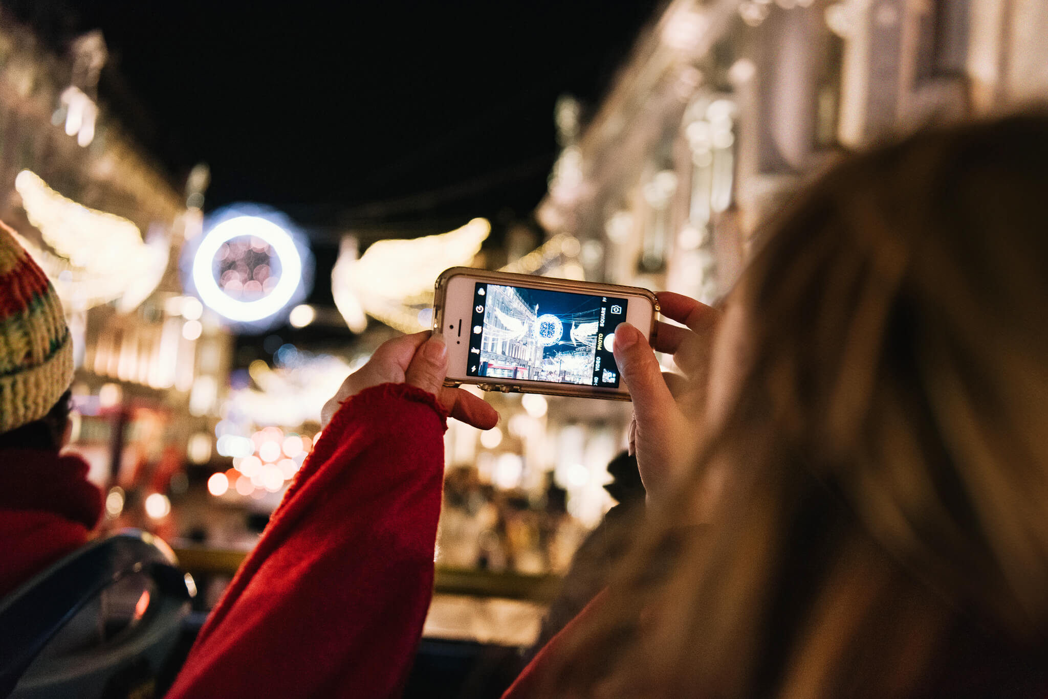 Vista trasera de una pasajera en el piso superior de un Tootbus, usando su teléfono inteligente para fotografiar las luces navideñas de Regent Street en Londres.