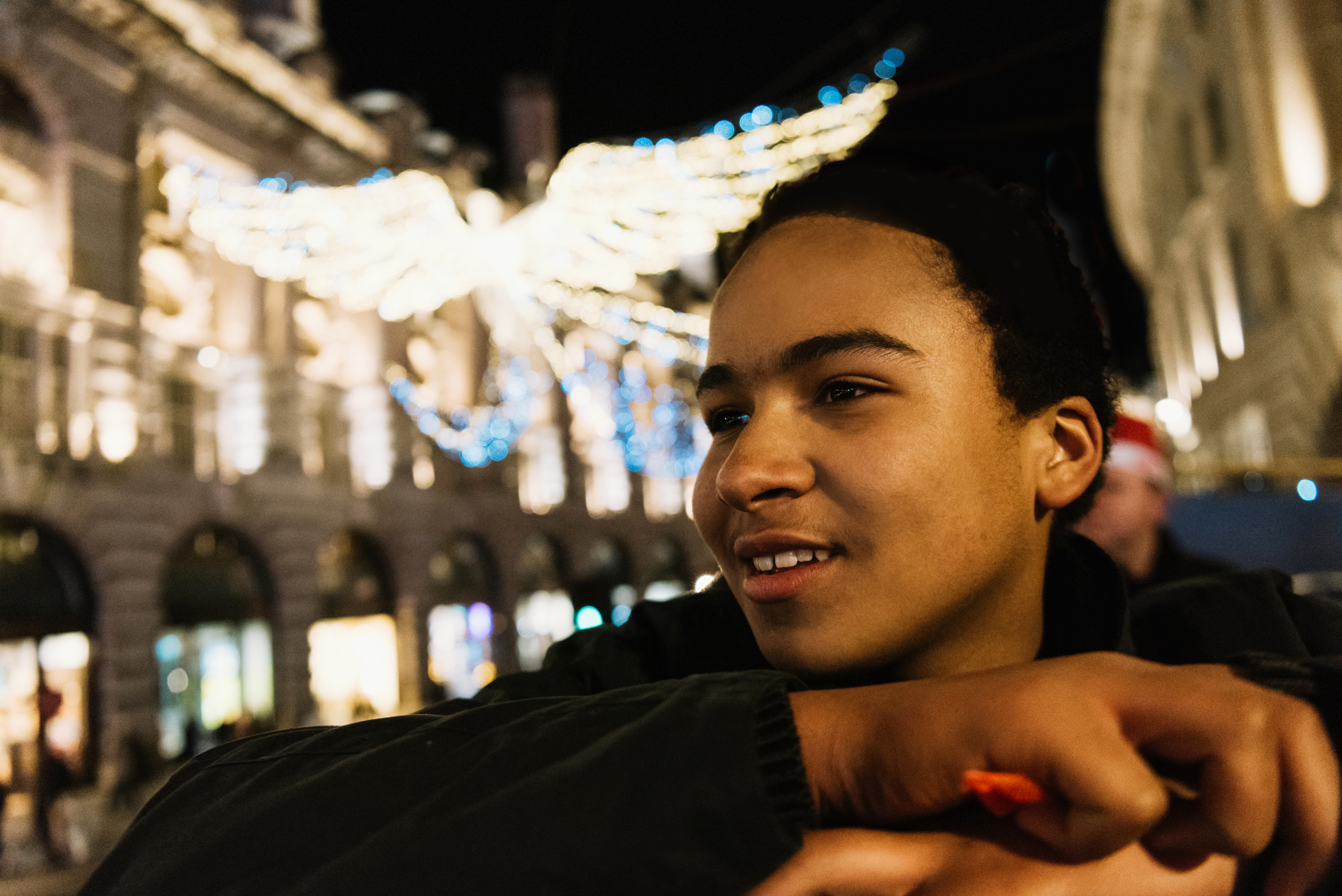 Un adolescente sonriente en el piso superior de un autobús descapotable en Londres, con hermosas luces navideñas en forma de ángel desenfocadas al fondo.
