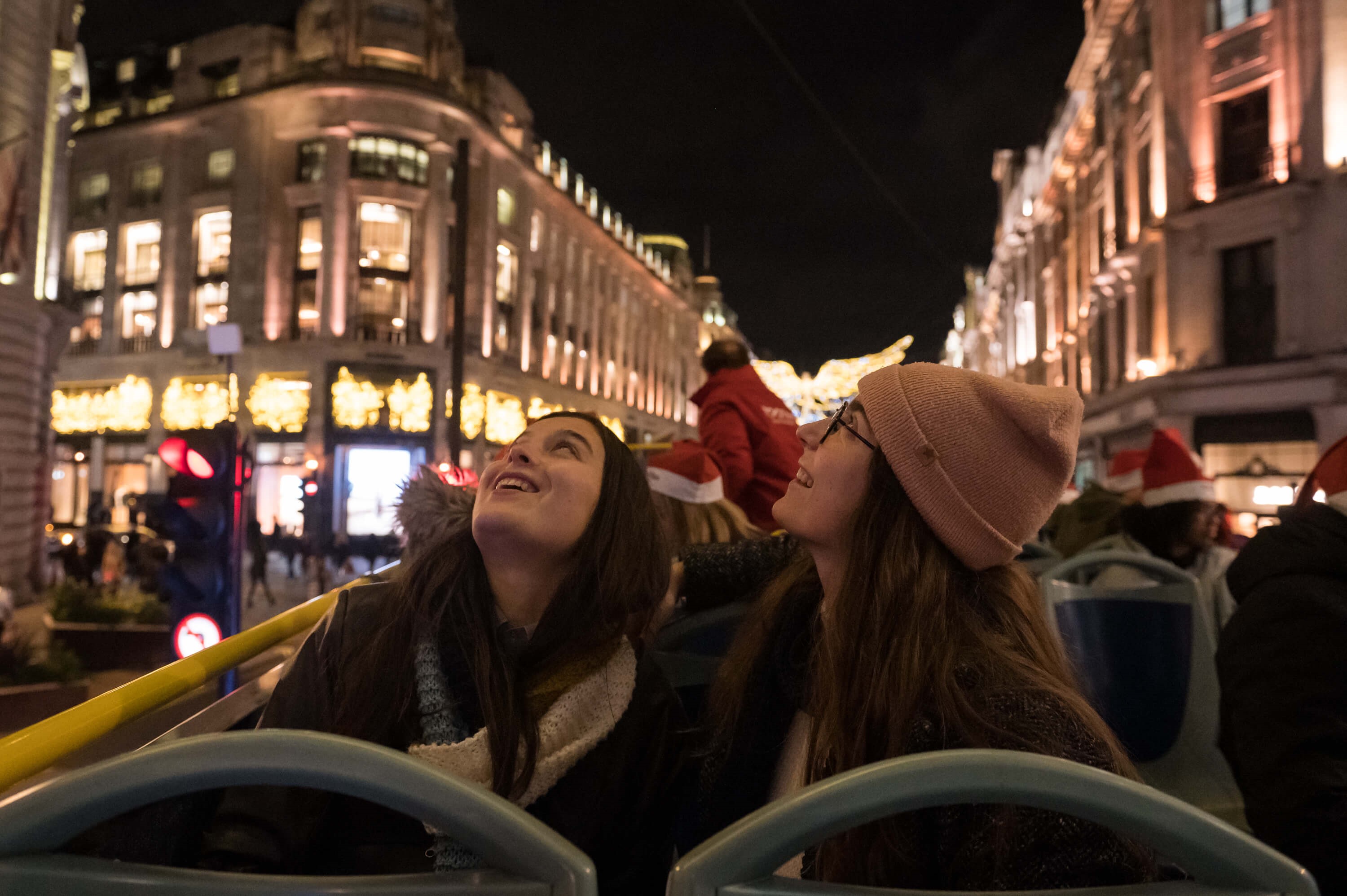 Dos jóvenes sonrientes con ropa de invierno en el piso superior de un autobús descapotable, maravilladas por las luces navideñas de Londres de noche.