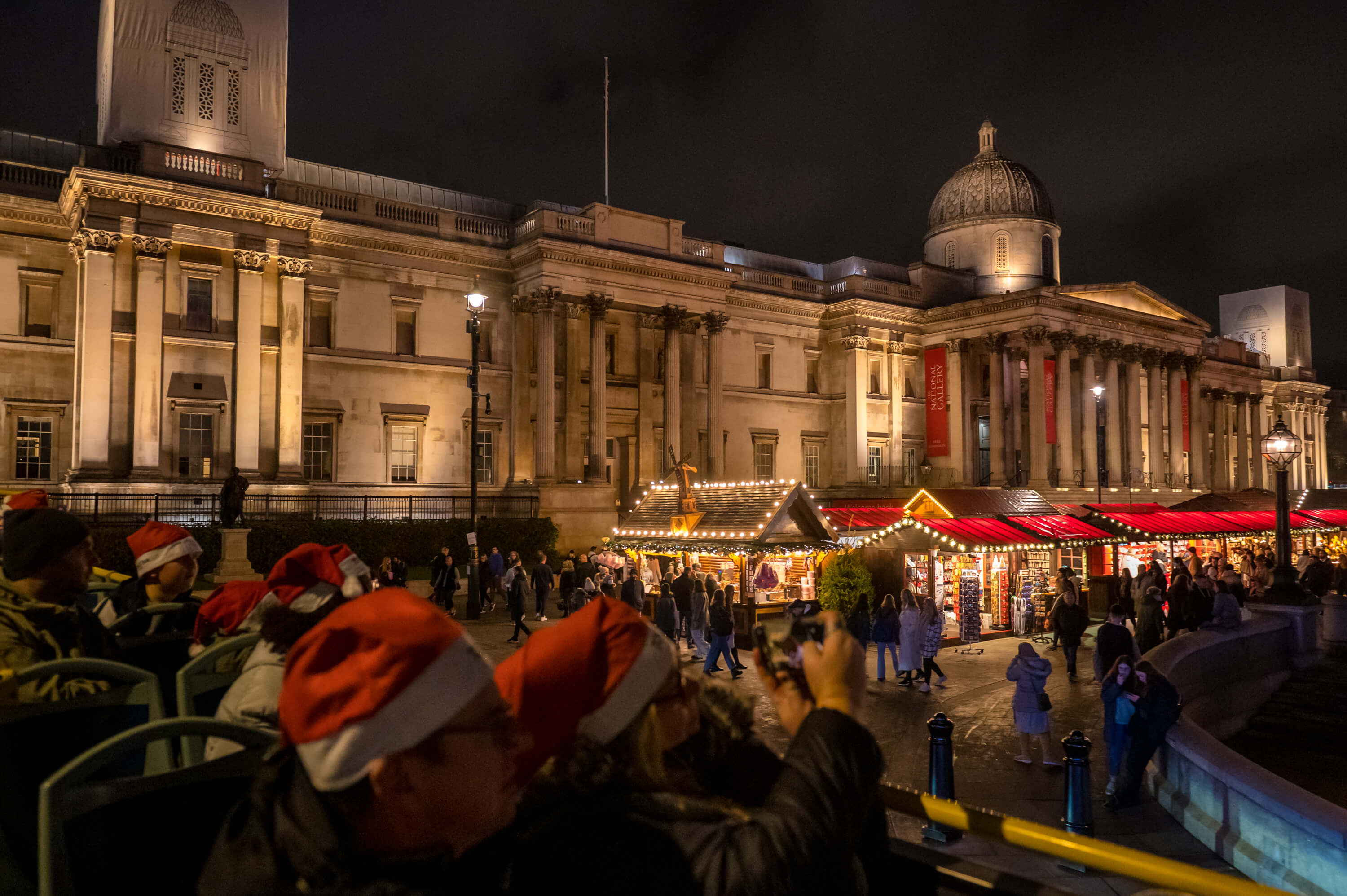 Pasajeros con gorros de Papá Noel en el piso superior de un Tootbus, admirando el mercado navideño iluminado frente a la National Gallery en Trafalgar Square.