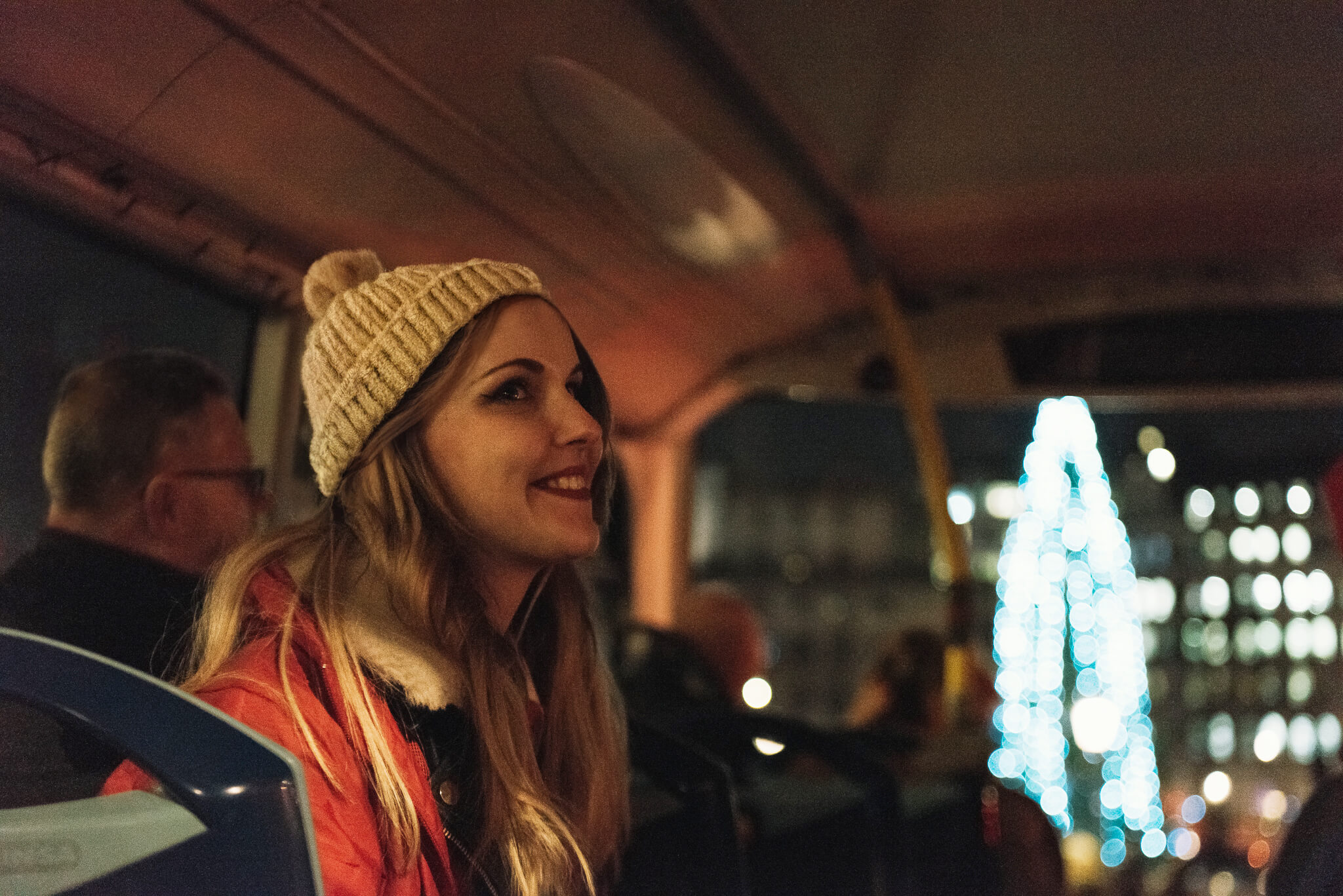 Una joven sonriente con un gorro de lana sentada en un autobús de dos pisos en Londres, con un gran árbol de Navidad iluminado de fondo desenfocado.