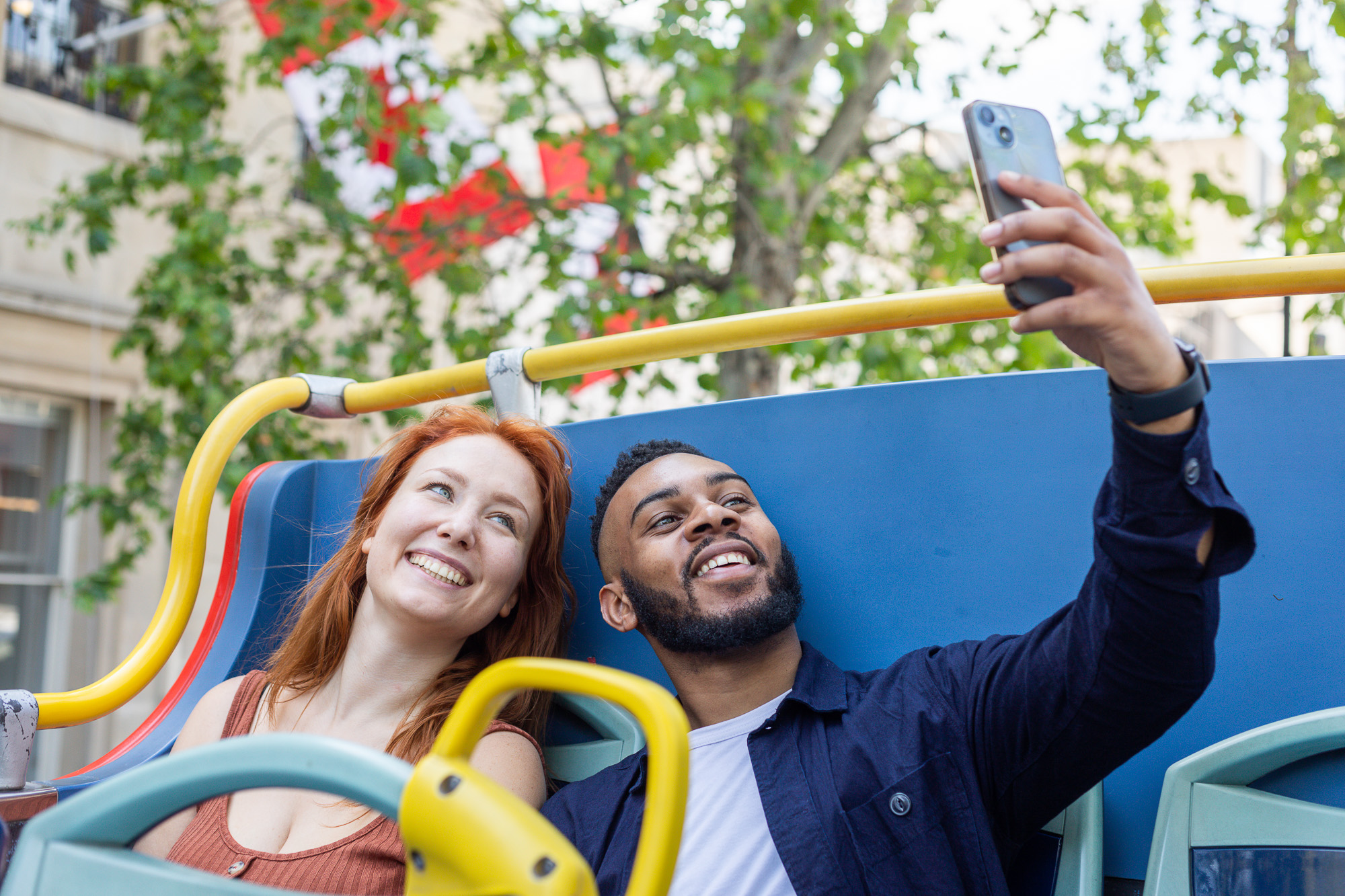 Un jeune couple souriant prenant un selfie avec un smartphone à l'étage supérieur d'un bus décapotable Tootbus à Londres.