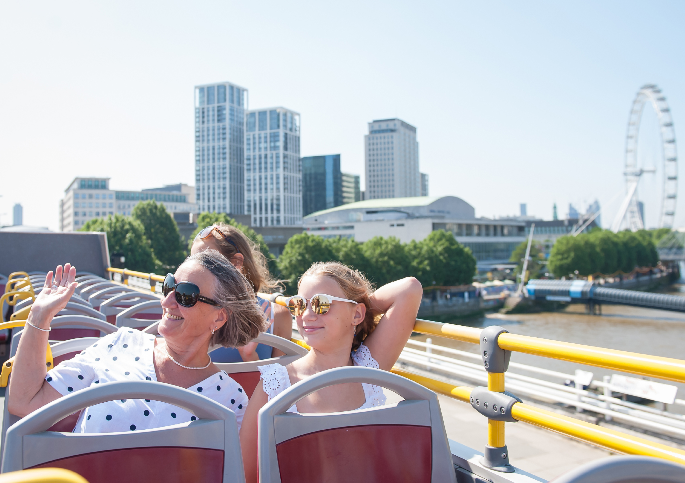 A mother and her daughter wearing sunglasses on the top deck of an open-top bus, smiling and waving with the London Eye Ferris wheel in the background in London.