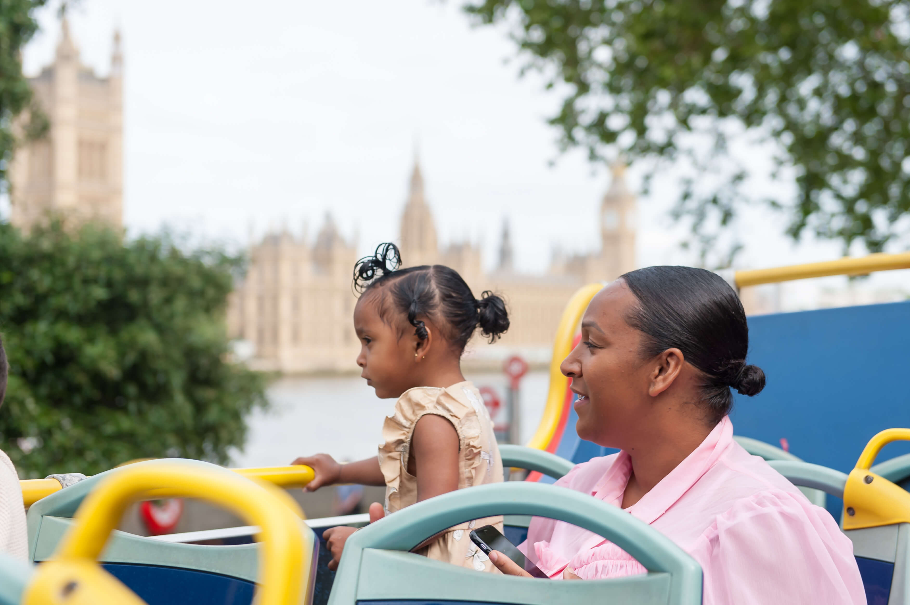 A woman and a young child sitting on the top deck of a Tootbus in London, looking at the Palace of Westminster across the River Thames.