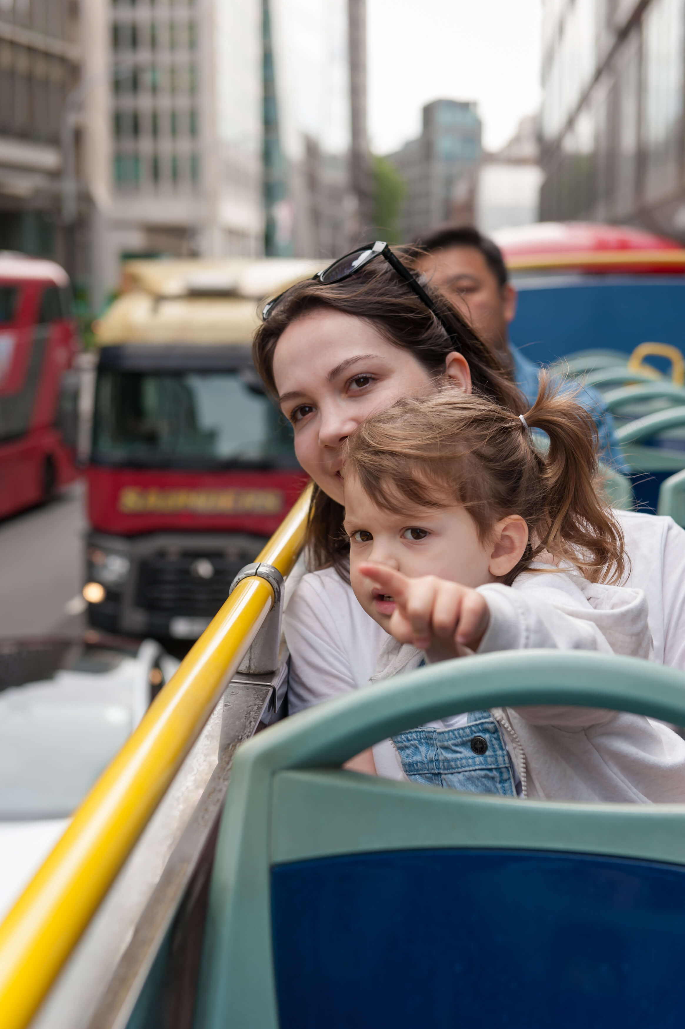 A smiling mother and her young daughter sitting on the top deck of an open-top bus in London, with the little girl pointing at a city landmark.