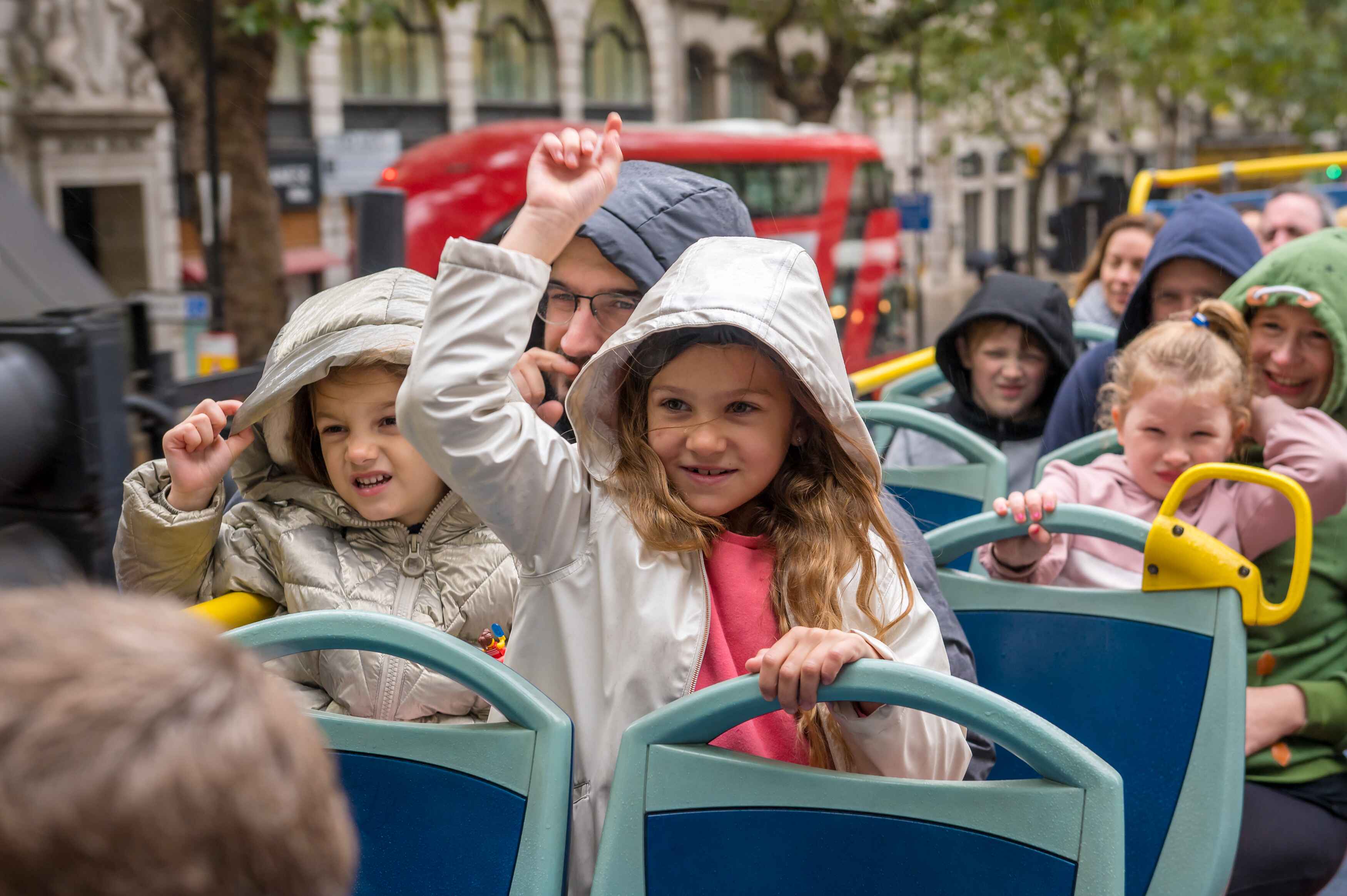 Enthusiastic children wearing hoods on the top deck of an open-top bus in London during a rainy day.