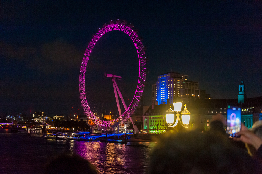 Vue nocturne de la grande roue du London Eye illuminée en rose à Londres, avec le reflet des lumières sur la Tamise et un passager prenant une photo.