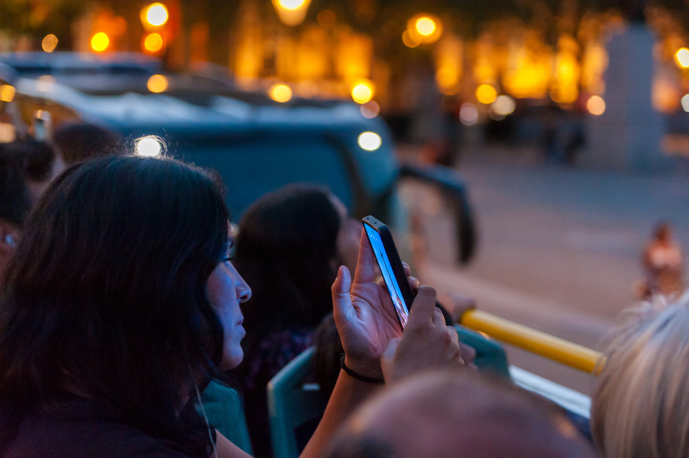 Profil d'une passagère à l'étage supérieur d'un bus décapotable Tootbus, utilisant son smartphone pour prendre une photo lors d'une visite nocturne de Londres.