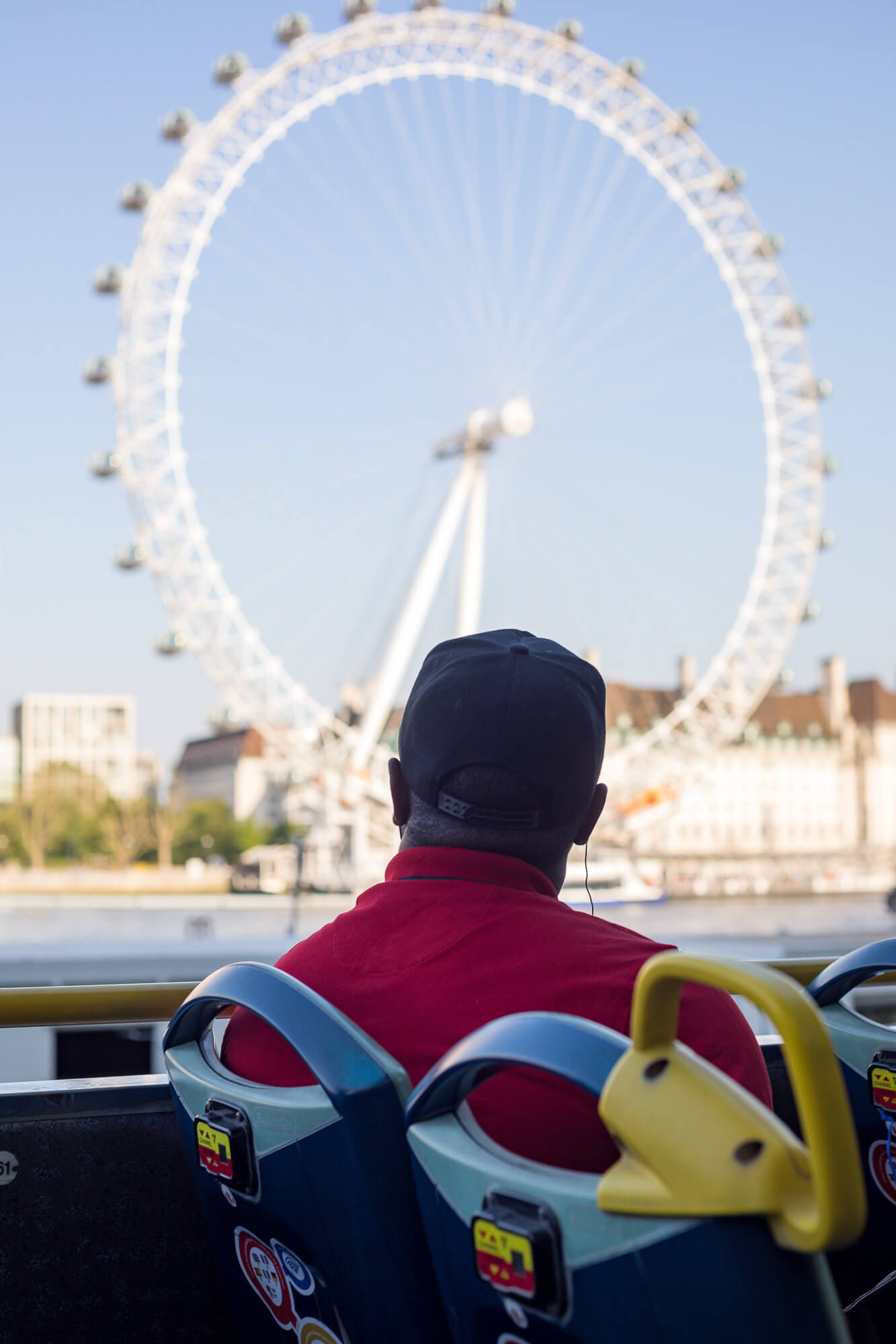 Vue de dos d'un passager portant une casquette noire et un t-shirt rouge à l'étage supérieur d'un bus décapotable, admirant la grande roue du London Eye à Londres.