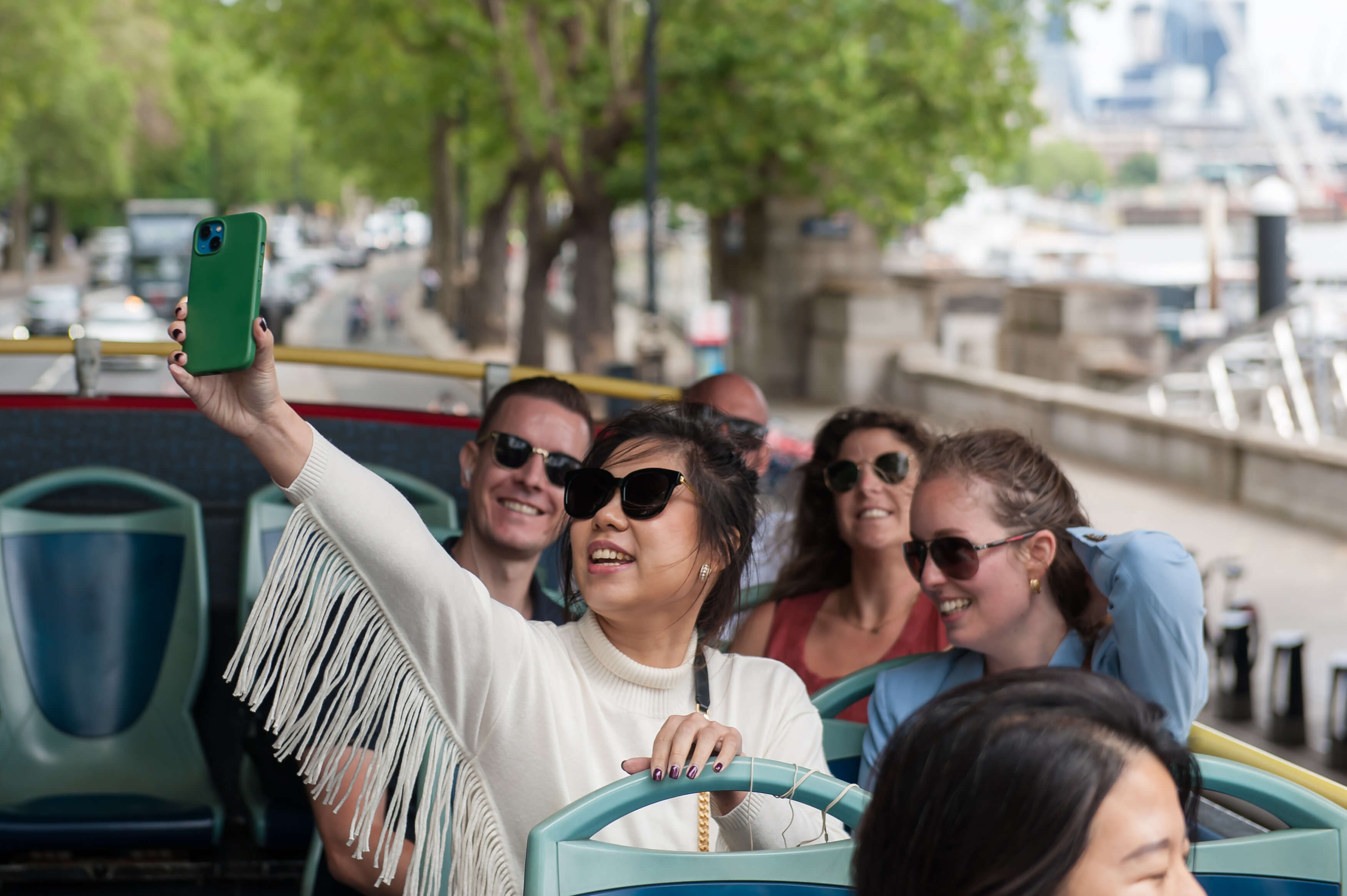 Un groupe de touristes souriants avec des lunettes de soleil prenant un selfie avec un smartphone à l'étage supérieur d'un bus décapotable Tootbus à Londres.