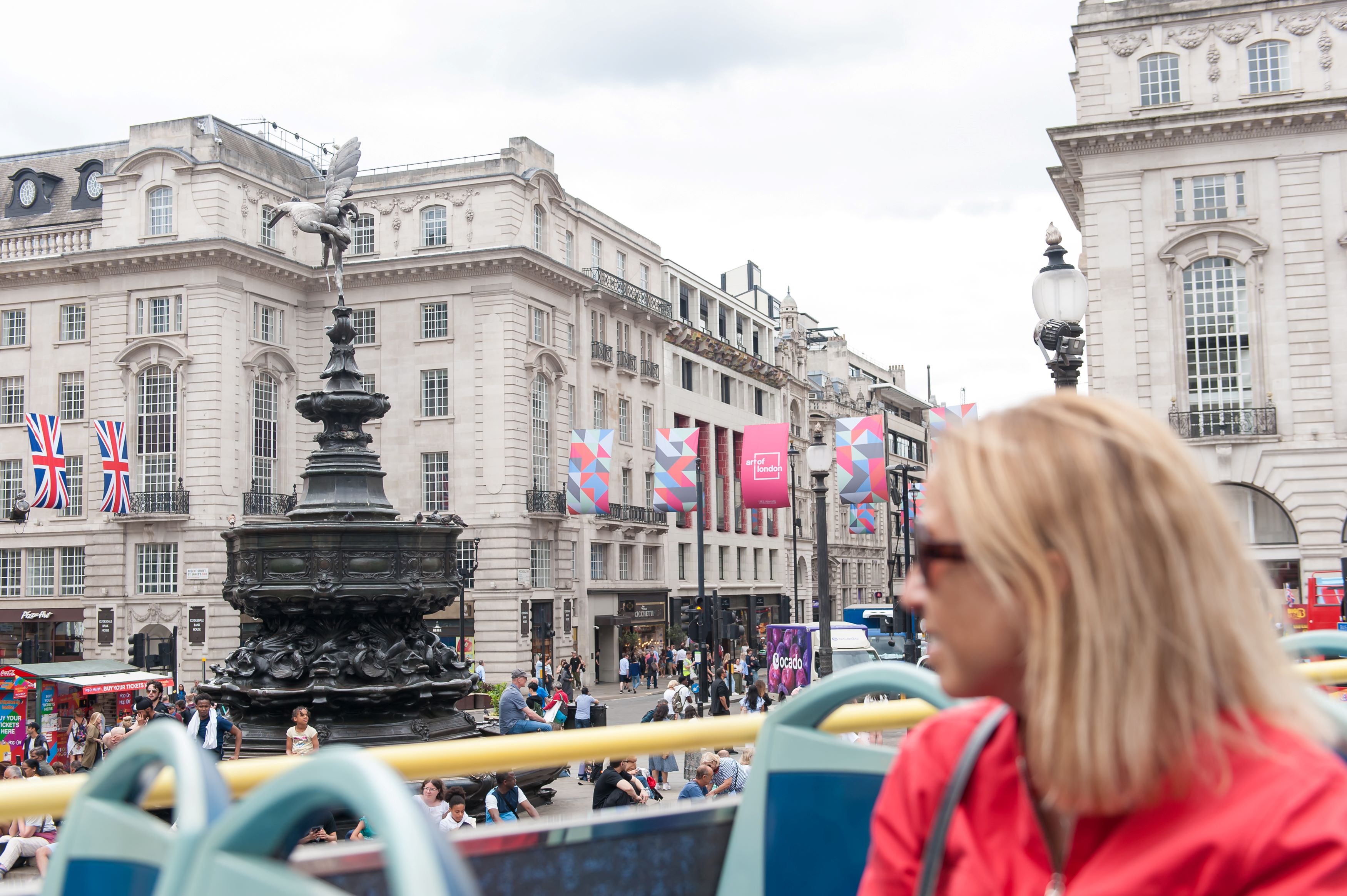 Une femme blonde avec des lunettes de soleil assise à l'étage supérieur d'un bus décapotable, passant devant la célèbre fontaine de Piccadilly Circus à Londres.