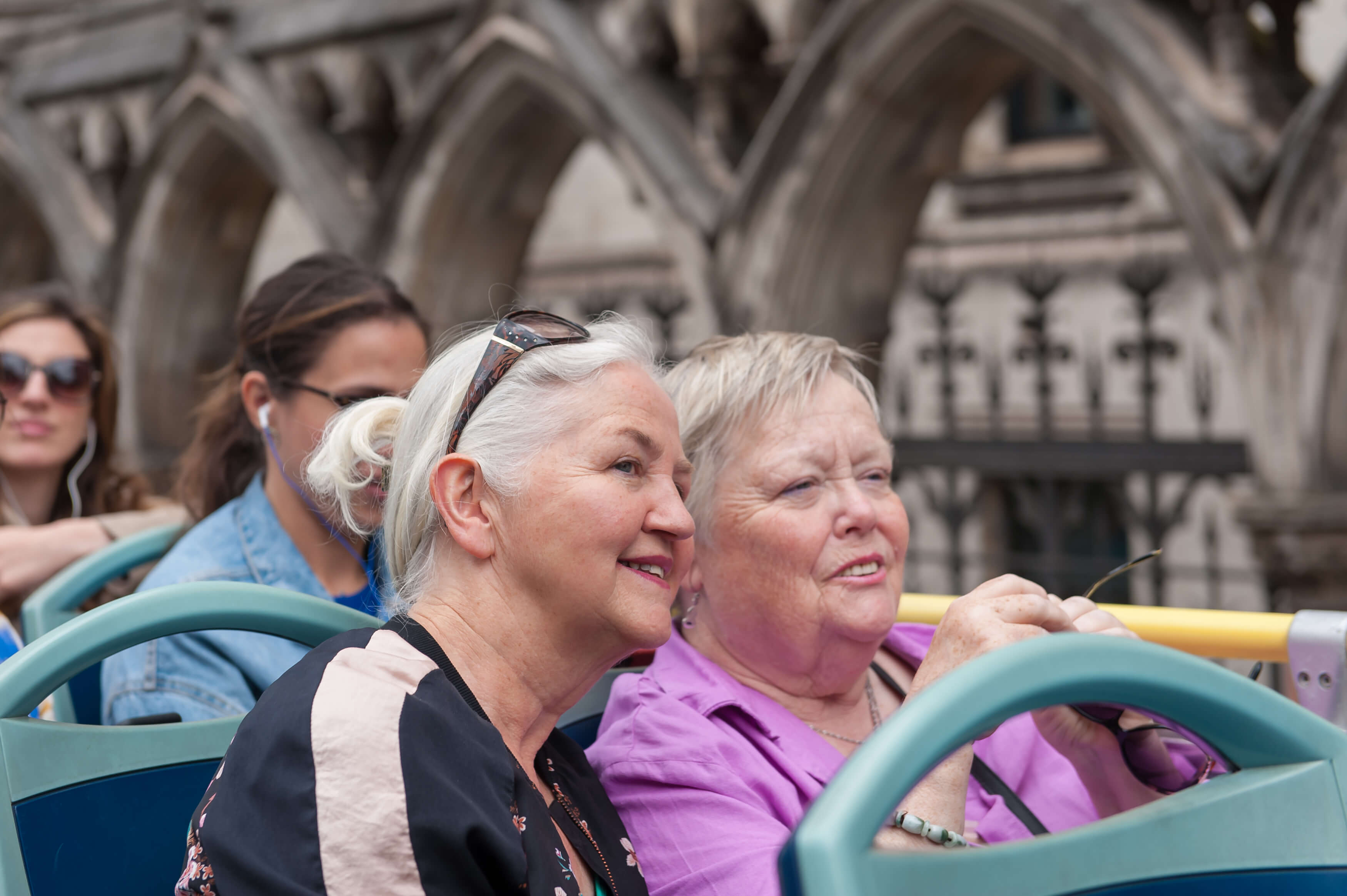 Deux femmes séniors souriantes assises à l'étage supérieur d'un bus Tootbus à Londres, admirant l'architecture gothique en pierre.