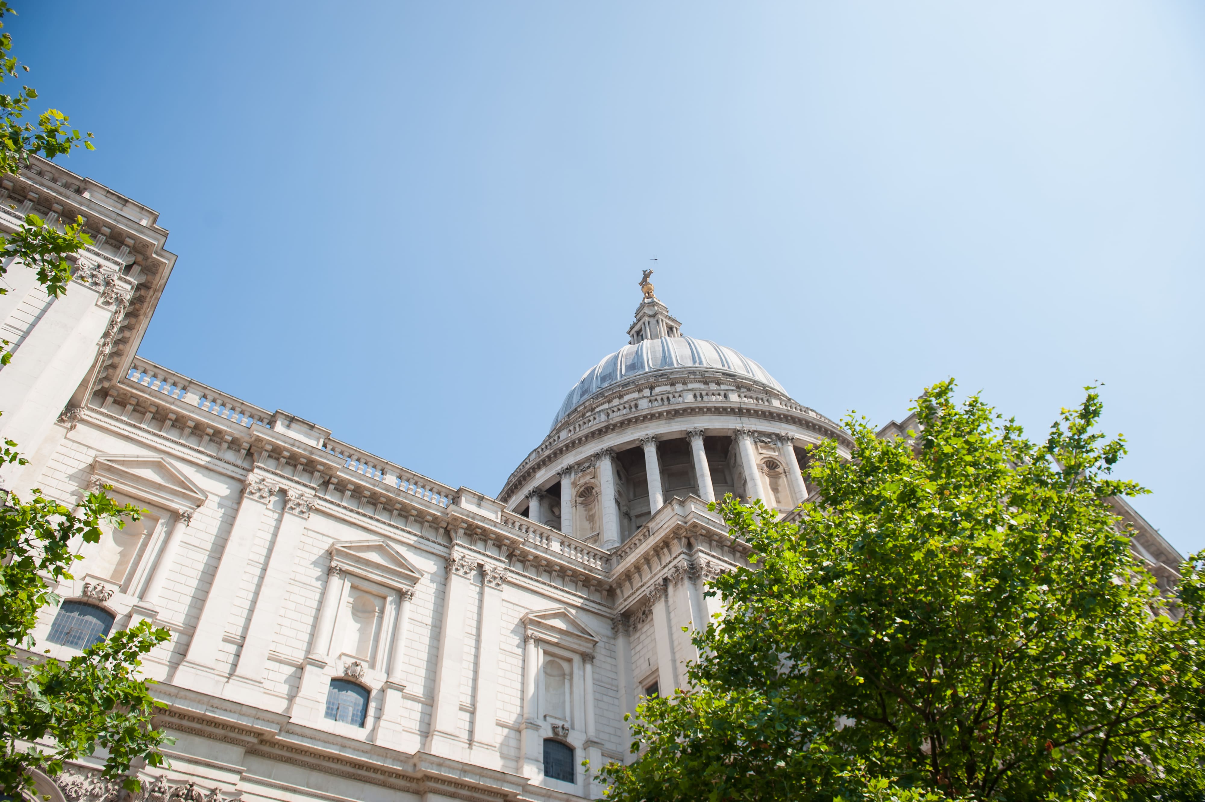 Low-angle view of St Paul's Cathedral in London, showing its white stone facade and majestic dome framed by green trees under a blue sky.