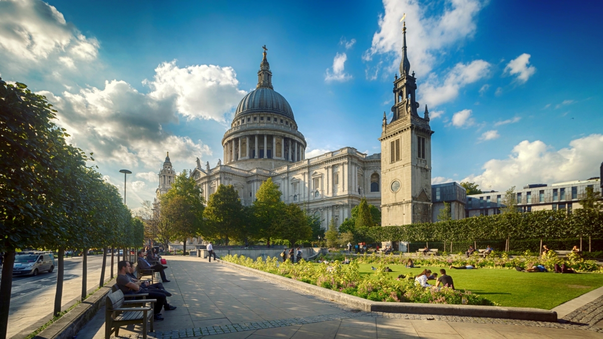 View of St Paul's Cathedral in London from an adjacent public garden, with people sitting on benches and the lawn on a sunny day.