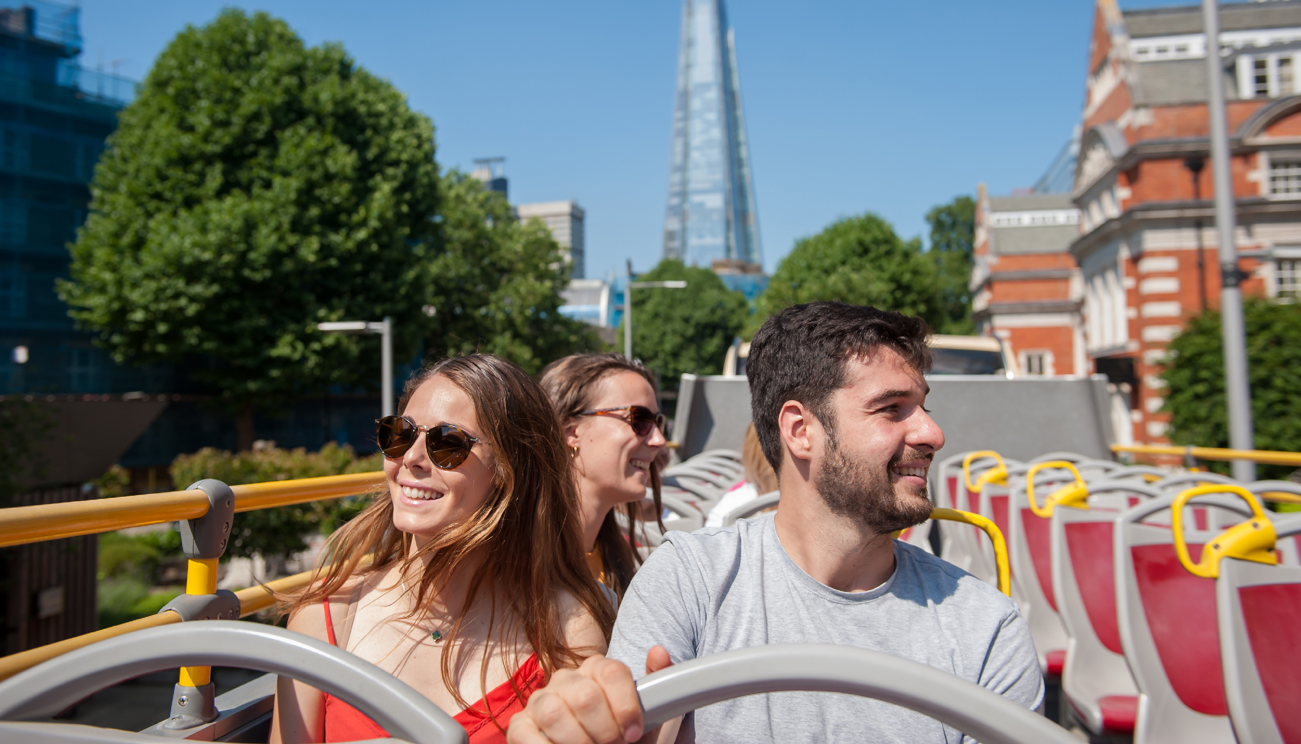 Trois jeunes touristes souriants assis à l'étage supérieur d'un bus décapotable à Londres, avec le gratte-ciel The Shard en arrière-plan.