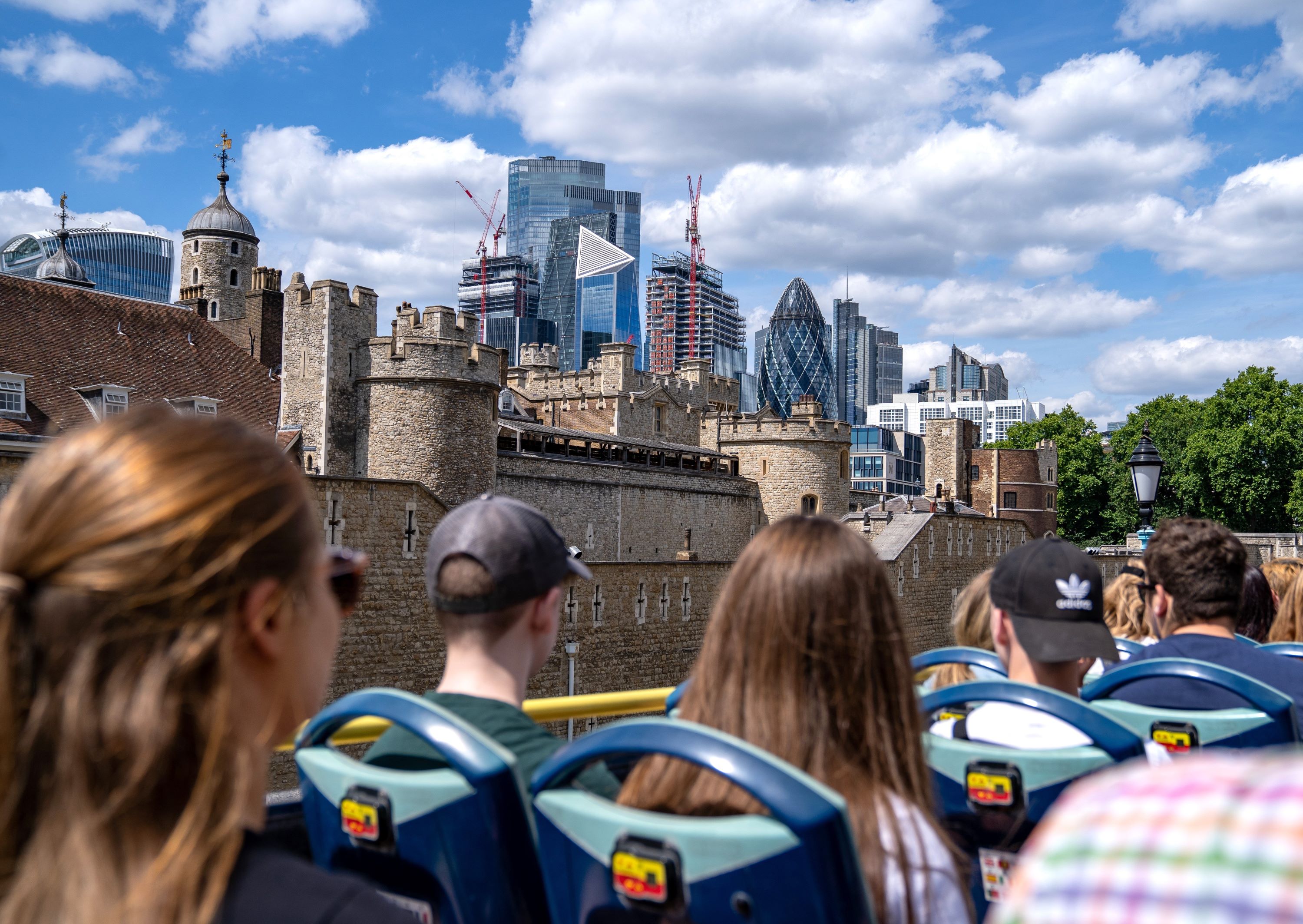 Vue de dos de passagers assis à l'étage supérieur d'un bus décapotable à Londres, admirant la forteresse historique de la Tour de Londres avec les gratte-ciels de la City en arrière-plan sous un ciel nuageux.