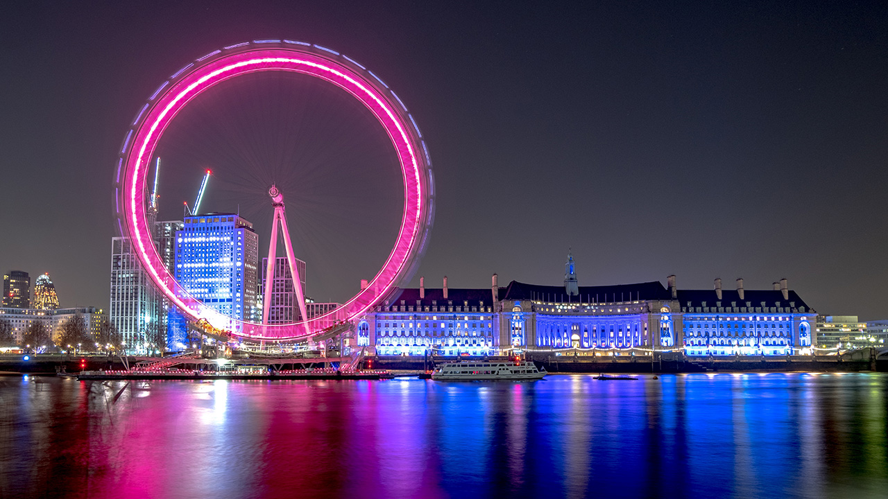 The London Eye Ferris wheel illuminated in bright pink and County Hall lit up in blue at night, reflecting on the River Thames in London.
