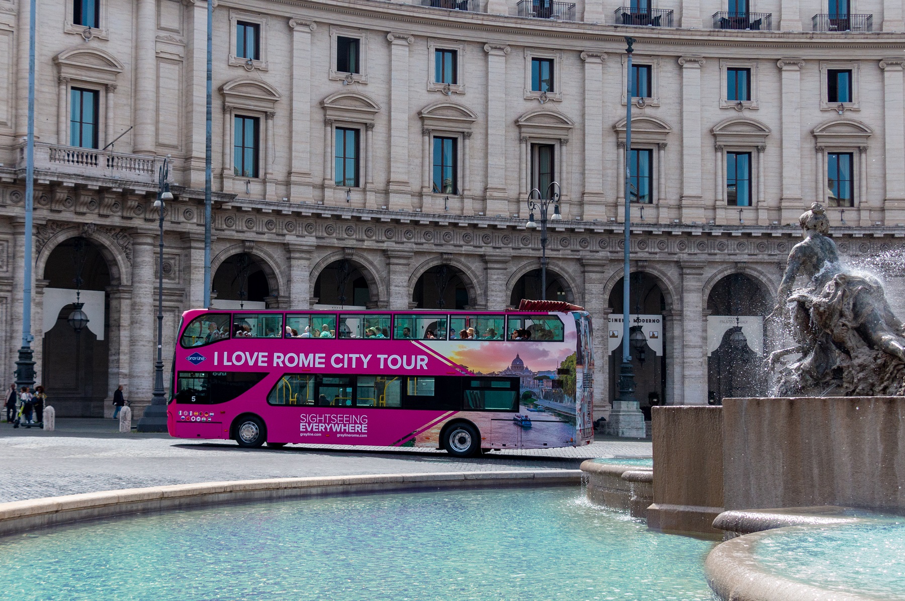 Pink « I Love Rome » hop-on hop-off bus driving across a plaza near a fountain in Rome.