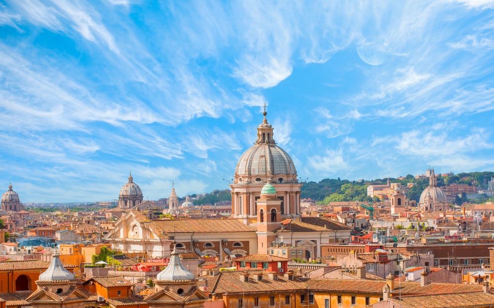 Panoramic view over the rooftops, domes, and churches of Rome under a cloudy sky.