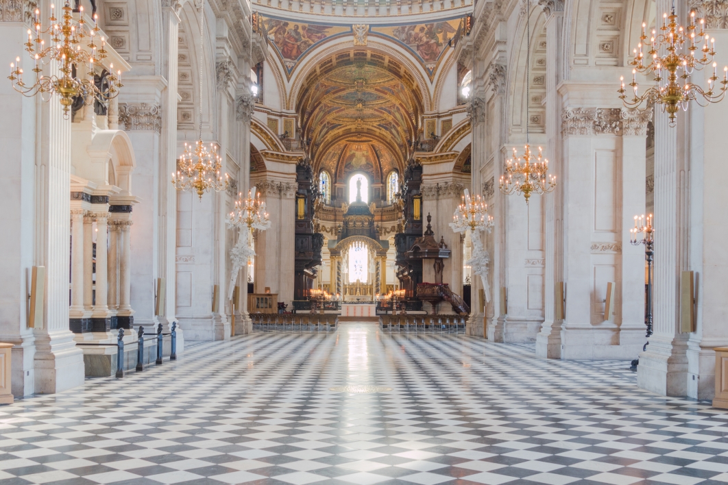 View of the majestic interior of St Paul's Cathedral in London, showing its checkered floor, ornate golden chandeliers, and nave leading to the altar.