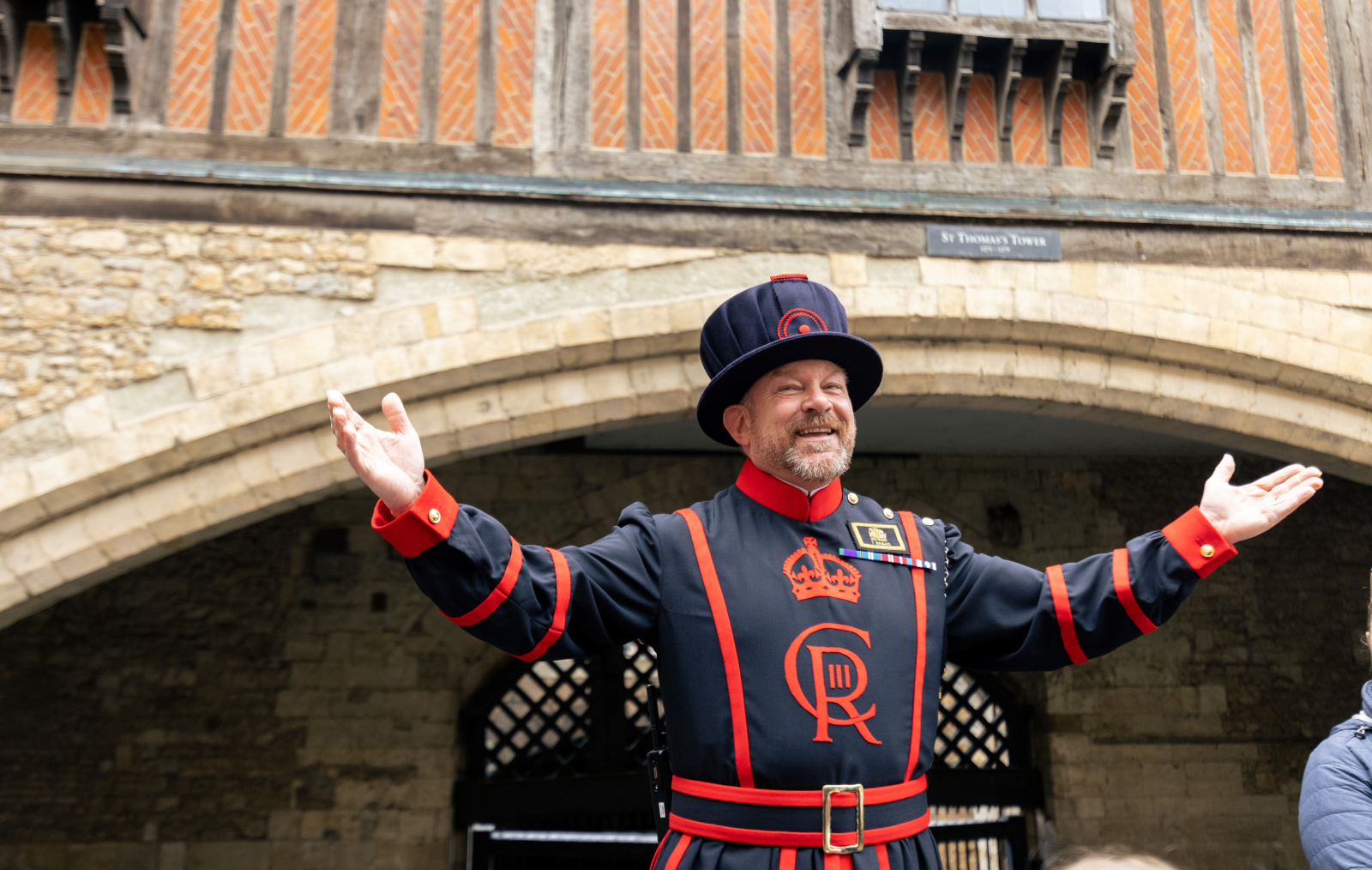 Un Yeoman Warder (Beefeater) sonriente con uniforme tradicional abriendo los brazos frente a la Torre de San Tomás en la Torre de Londres.