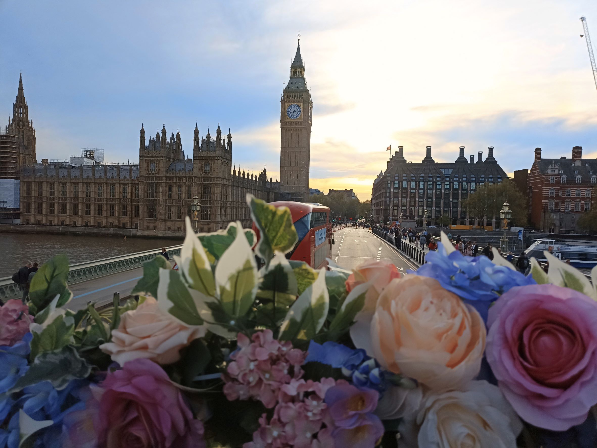 View from the flower-decorated top deck of a bus crossing Westminster Bridge, featuring Big Ben and the Houses of Parliament in the background.