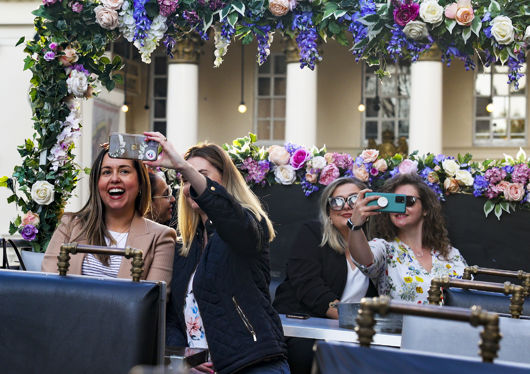 Group of smiling friends taking selfies on the flower-decorated top deck of a bus during a private tour in London.