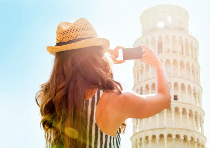 Tourist from behind wearing a straw hat and taking a photo of the famous leaning Tower of Pisa with a smartphone under bright sunlight.