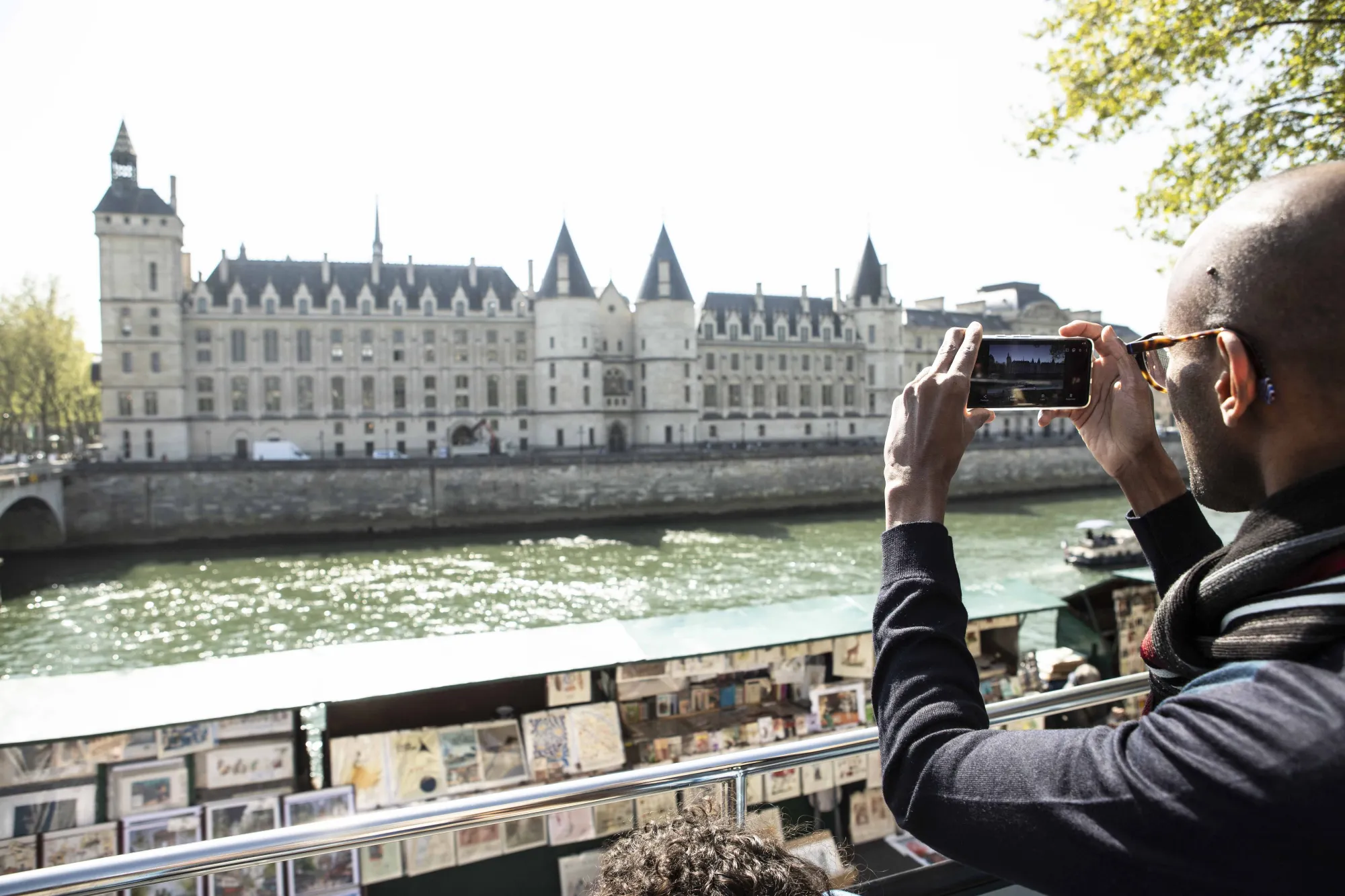 Tourist in profile taking a photo with a smartphone from the upper deck of a bus facing the Conciergerie and booksellers along the Seine river.