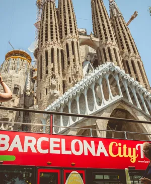 Tourists on the upper deck of a red Barcelona City Tour bus taking photos of the Sagrada Família basilica in Barcelona.