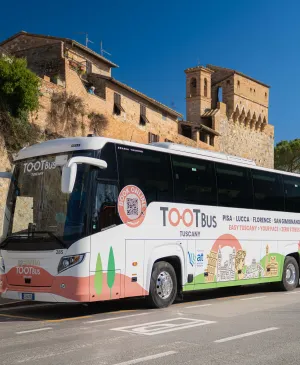 White Tootbus Tuscany tour coach parked along a road near an ancient medieval stone wall under a clear blue sky in Tuscany.